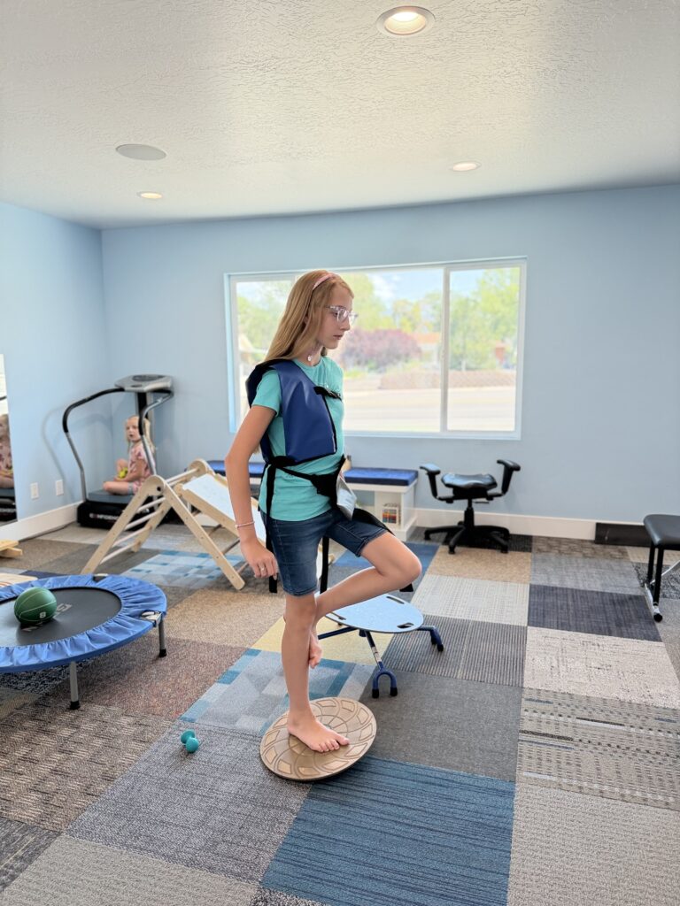 A child balancing on a wobble board in a weighted vest during a postural session at Kids Only Chiropractic